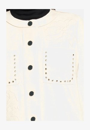 Witte jas met een ronde kraag, twee voorzakken versierd met studs, en een reliëf bloemmotief aan de linkerzijde. Zwarte knopen aanwezig.