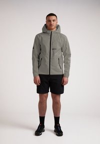 Giacca impermeabile grigia chiara con cappuccio, zip frontale e due tasche laterali, abbinata a pantaloni corti neri e scarpe. Design minimal.