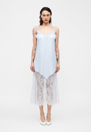 Femme aux cheveux foncés portant une robe bleu clair sans manches avec un ourlet en dentelle, debout pieds nus devant un fond blanc uni.