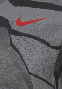 Camiseta atlética gris con patrones abstractos en negro y gris más oscuro. Presenta un prominente logo de swoosh rojo de Nike. Textura de tela suave.