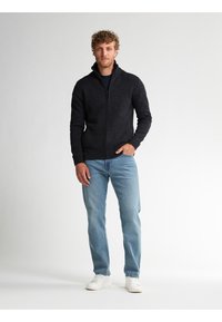 Mörkgrå zip-up tröja med ribbat krage och ärmslut, i kombination med ljusblå raka jeans och vita sneakers. Neutral bakgrund.
