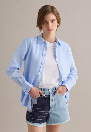 Junge Person mit kurzen Haaren trägt ein hellblaues, geknöpftes Hemd über einem weißen T-Shirt und Jeansshorts mit navyfarbenem Patchwork, Hände in den Taschen.