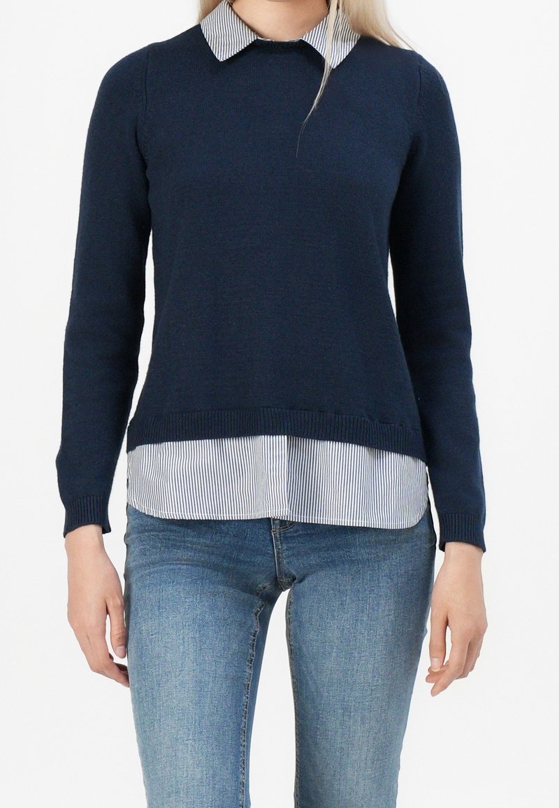 Femme portant un pull bleu marine par-dessus une chemise à col rayée bleu et blanc et un jean bleu clair, debout devant un fond blanc.