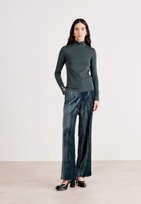 Dark green fitted turtleneck top paired with pleated wide-leg trousers, showcasing a textured fabric and a smooth silhouette. Black platform shoes.