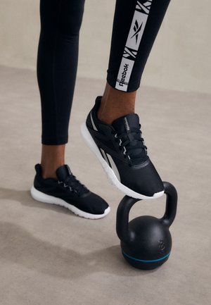 Personne portant des leggings Reebok noirs et des baskets noires marche sur une kettlebell noire sur un sol gris clair.