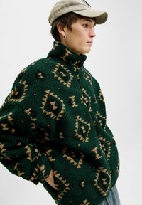 Grüne Fleecejacke mit hohem Kragen, die geometrische tanfarbene Muster aufweist. Hergestellt aus weichem Stoff, verfügt sie über einen Reißverschluss und lange Ärmel.