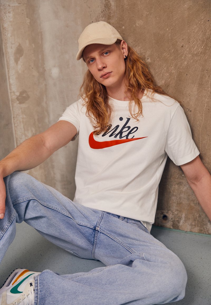 T-shirt bianco in cotone con logo Nike rosso e nero, abbinato a jeans di denim blu chiaro. La persona indossa un cappello beige e sneakers bianche con accenti di colore.