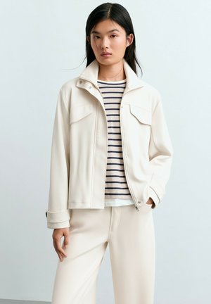 Femme portant une veste crème par-dessus une chemise blanche à rayures bleu marine avec un pantalon crème assorti, debout devant un fond clair uni.
