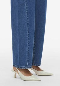 Bāli beige lakotas ādas puses ar norādītu purngalu un zemu bloku papēdi, apvienotas ar ziliem platajiem džinsu šortiem.