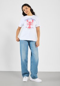 Hvid bomulds T-shirt med et rødt hummer grafik og blå tekst, der siger "Maine Beach." Sammen med lyseblå vide jeans og hvide sneakers.