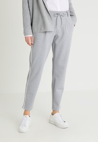 Hellgraue Jogginghose aus weichem Stoff mit einem Kordelzugbund und weißen Seitenstreifen. Kombiniert mit weißen Turnschuhen und einer grauen Strickjacke.