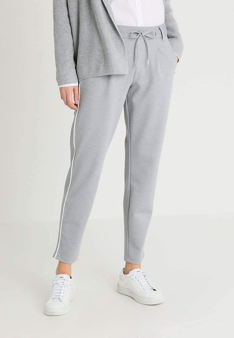 Hellgraue Jogginghose aus weichem Stoff mit einem Kordelzugbund und weißen Seitenstreifen. Kombiniert mit weißen Turnschuhen und einer grauen Strickjacke.