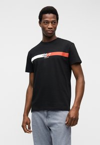 Άνδρας που φοράει μαύρο t-shirt Tommy Jeans με λευκή και κόκκινη ρίγα, συνδυασμένο με ανοιχτόχρωμο τζιν, στέκεται μπροστά σε απλό ανοιχτό φόντο.