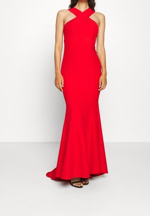 Kobieta ubrana w dopasowaną, czerwoną suknię sięgającą podłogi z krzyżującym się halterem na szyi i czarne sandały z odkrytymi palcami, stoi na tle jednolitego tła.