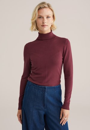 Burgunderfarbener Rollkragenpullover aus einem weichen, dehnbaren Material mit langen Ärmeln, kombiniert mit lockeren blauen Jeanshosen, die über einen elastischen Bund verfügen.