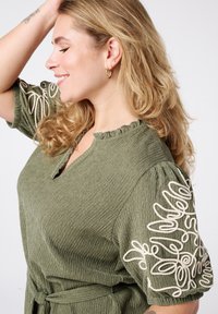 Olijfgroen, gestructureerde jurk met een gerimpelde halslijn en korte pofmouwen met witte geborduurde details op de schouders.