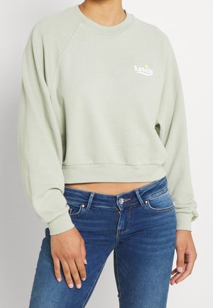 Personne portant un sweat court Levi's vert clair et un jean bleu, debout avec une main reposant sur la cuisse.