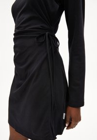 Schwarzes Wickelkleid mit langen Ärmeln, weich strukturiertem Stoff, verstellbarem Taillenband, seitlichem Schlitz und kleinem Markenschild am Saum.