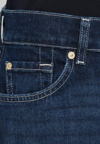 Calças jeans de ganga azul escuro com bolso frontal, fecho com botão dourado e costura em contraste ao longo do cós e das bordas dos bolsos.