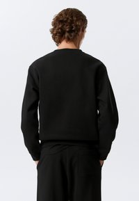 Sweat-shirt noir à col rond, fabriqué en tissu doux. Présente une coupe décontractée et des poignets côtelés. La vue de dos montre une texture lisse et des lignes épurées.