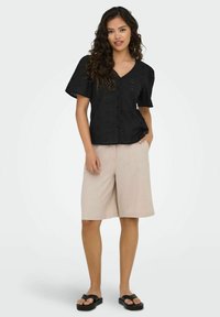 Schwarze Bluse mit kurzen Ärmeln, durchbrochenen Details und V-Ausschnitt, kombiniert mit beigen knielangen Shorts und schwarzen Riemchensandalen.