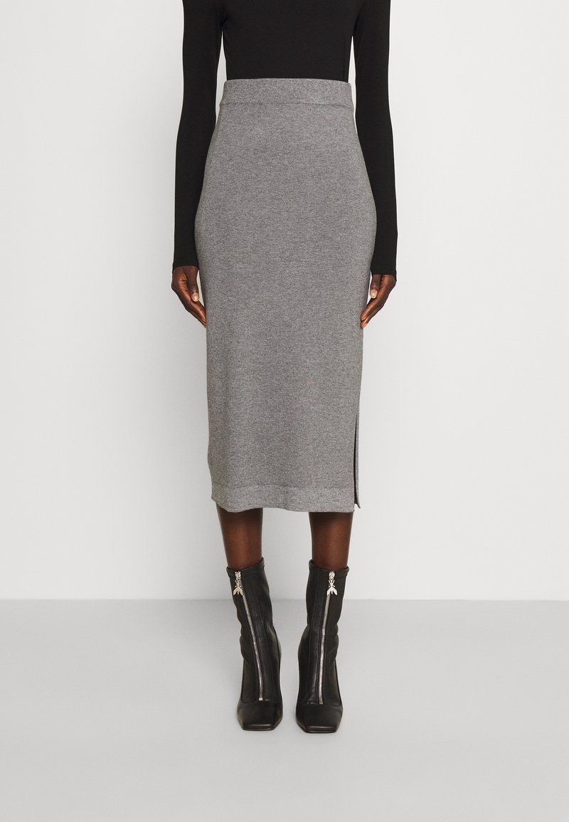 Femme portant une jupe midi grise taille haute avec une fente latérale, un top noir à manches longues et des bottines noires à talons avec fermetures éclair à l'avant.