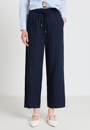 Donna che indossa pantaloni blu navy a gamba larga con vita regolabile con coulisse, abbinati a ballerine bianche a punta e camicia azzurra a righe annodata.