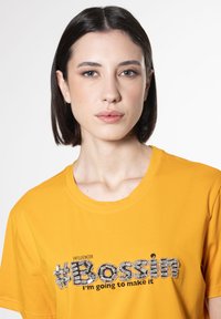 Gul bomulds t-shirt har en rund halsudskæring og korte ærmer. Centreret tekst inkluderer "#BOSSin" i strukturerede bogstaver og "Jeg vil få det til at ske."
