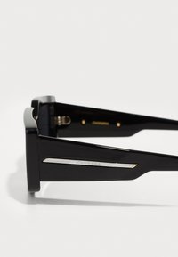 Ochelari de soare negri cu un design elegant, având rame lucioase, formă unghiulară, accente metalice și un logo argintiu pe tâmplă.