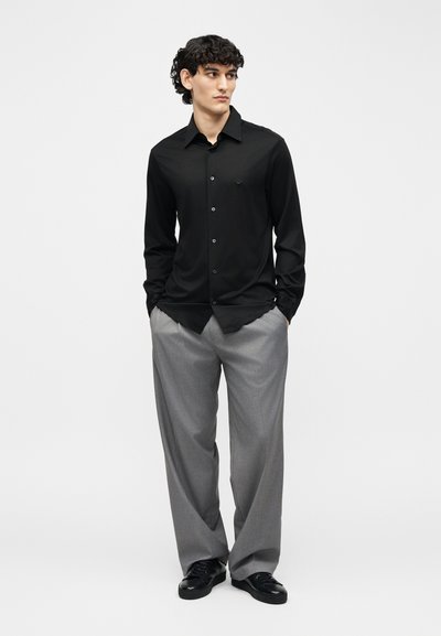Νέος άνδρας που φοράει μαύρο πουκάμισο με κουμπιά και γκρίζα φαρδιά παντελόνια, στέκεται με τα χέρια στις τσέπες, κοιτώντας προς το πλάι πάνω σε ένα απλό φόντο.