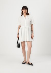 Rochie albă din in cu mâneci scurte, guler și talie strânsă. Poartă încălțări negre tip slip-on și o poșetă mică neagră.