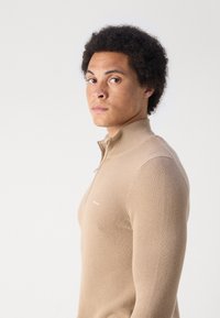 Maglione beige con zip e collo a lupetto, caratterizzato da un design a maglia testurizzata. Presenta un logo discreto sul petto e una silhouette aderente.