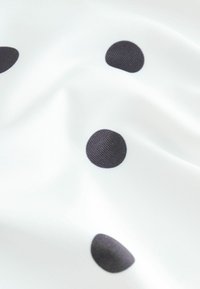 Vitt tyg med stora svarta prickar, som visar en slät textur med en lätt glans. Mönstret är jämnt fördelat över materialet.