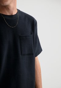 Námorně modré tričko s krátkým rukávem vyrobené z fleecového materiálu texturované na povrchu, vybavené kapsou na prsou a vroubkovaným kulatým výstřihem. Viditelný zlatý řetízek jako detail.