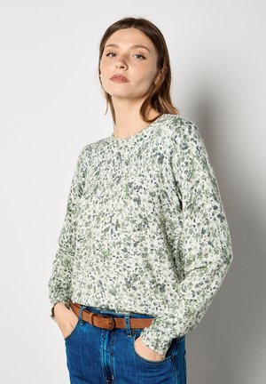 Groen-witte top met lange mouwen en bloemmotief, voorzien van een ronde halslijn en geribbelde manchetten, gecombineerd met blauwe high-waisted jeans en een bruine riem.