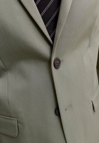 Κοντινή λήψη ενός πράσινου σακακιού με δύο σκούρα κουμπιά πάνω σε μαύρη γραβάτα με λευκές διαγώνιες ρίγες.