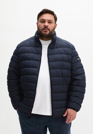 Mann, der eine marineblaue gesteppte Jacke über einem weißen T-Shirt trägt, steht vor einem einfarbigen weißen Hintergrund und schaut nach vorne.