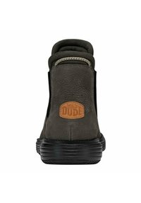 Botín de cuero marrón oscuro con suela de goma negra, cuello acolchado y un parche de cuero beige etiquetado "HEY DUDE" en la parte trasera.