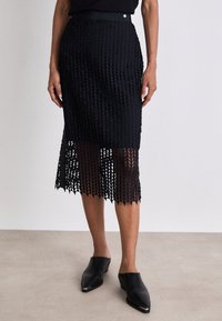 Zwarte, knielange rok met een kantachtige, geometrische patroon. Beschikt over een band rondom de taille en een golvende zoom. Draagt met zwarte instapschoenen.
