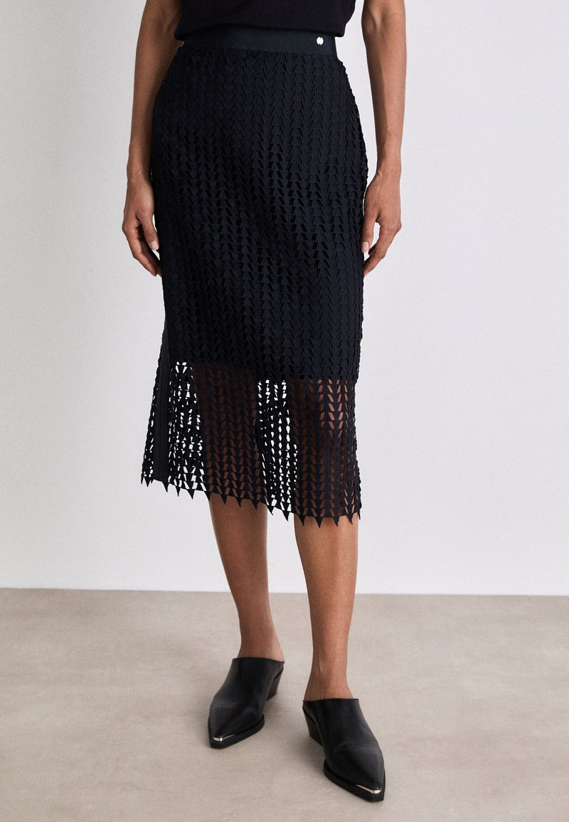 Zwarte, knielange rok met een kantachtige, geometrische patroon. Beschikt over een band rondom de taille en een golvende zoom. Draagt met zwarte instapschoenen.
