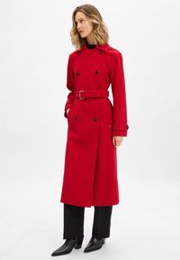 Cappotto rosso doppiopetto con cintura in vita, dotato di bottoni neri, tasche laterali e una linea dritta, indossato sopra pantaloni neri e stivali con tacco.