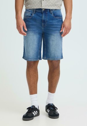 Mann iført denimshorts i mellomvask, hvite crew-sokker og svarte joggesko med hvite striper, stående mot en ensfarget bakgrunn.