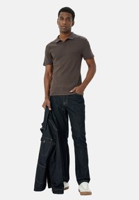 Brun ribbet polo-skjorte, mørkeblå jeans, holder en sort jakke. Modellen har kort hår, hvide sneakers og en afslappet holdning.