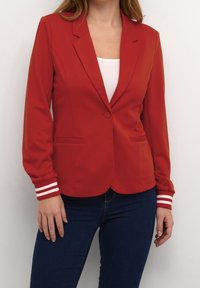 Rød tætsiddende blazer med notch-krave, enkelt knaplukning og to forlommer. Manchetter med hvide stribedetaljer. Glat stof.