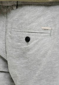 Κοντινό πλάνο σε ανοιχτό γκρι παντελόνι από ύφασμα με κουμπωτή πίσω τσέπη και μια μικρή ετικέτα "Jack & Jones" πάνω από την τσέπη.