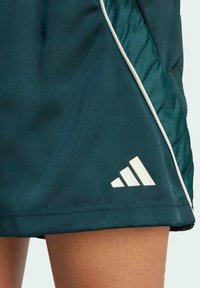 Tmavě zelená sportovní sukně vyrobená z hladkého, lehkého materiálu. Má bílé lemování a bílou logo Adidas na lemu.