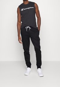 Svart ärmlös Champion-top med vit logotyp, kombinerad med svarta cargo-byxor med sidofickor och dragsko. Bärs med vita sneakers.