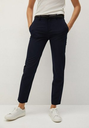 Persoon draagt slanke marineblauwe broek met zwarte riem, witte tanktop en witte sneakers, handen in de zakken, staand tegen een effen achtergrond.