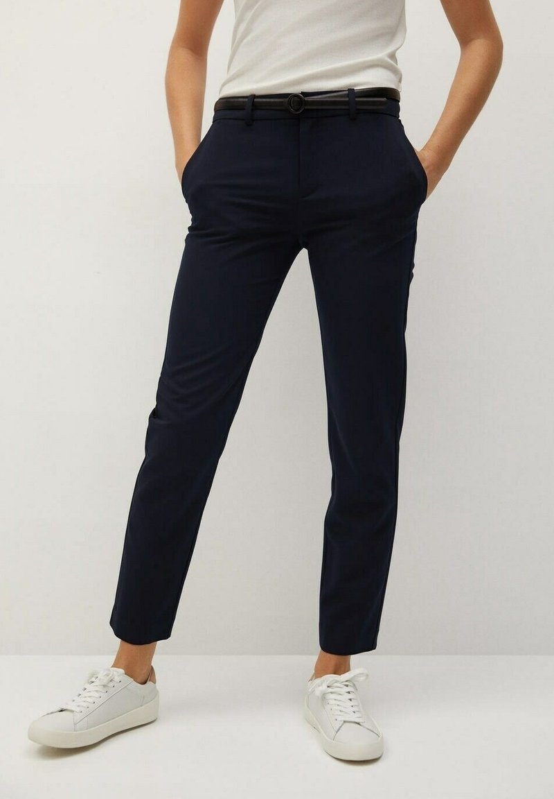 Personne portant un pantalon bleu marine ajusté avec ceinture noire, débardeur blanc et baskets blanches, mains dans les poches, debout devant un fond uni.