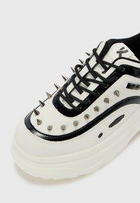 Deportivas blancas y negras con suela gruesa, detalles de charol y tachuelas metálicas en la parte superior. Tiene cordones negros y una textura suave.